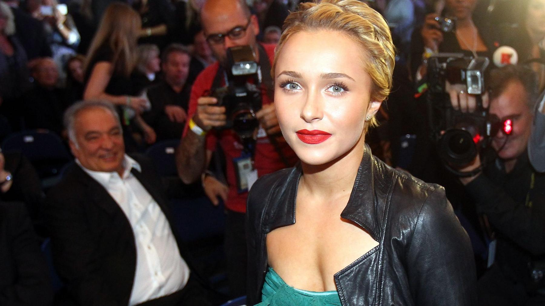 Hayden Panettiere surrounded by photographers she has her hair tied back and wearing red lipstick and a greendress with a black leather jacket over the top