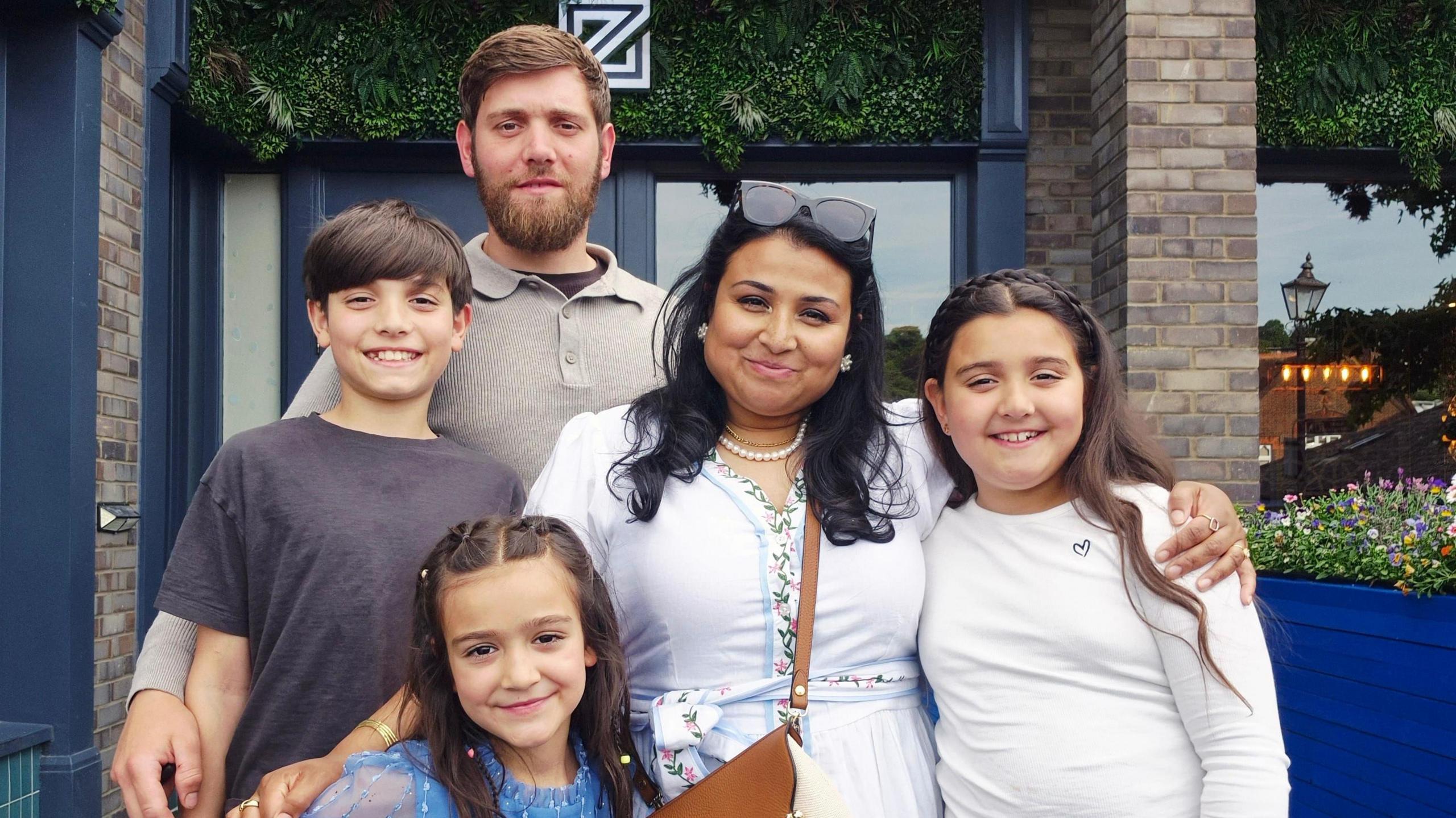A family photo, a man with brown hair and beard and a woman with dark hair and sunglasses on her head smiling with their three children, all have brown hair.