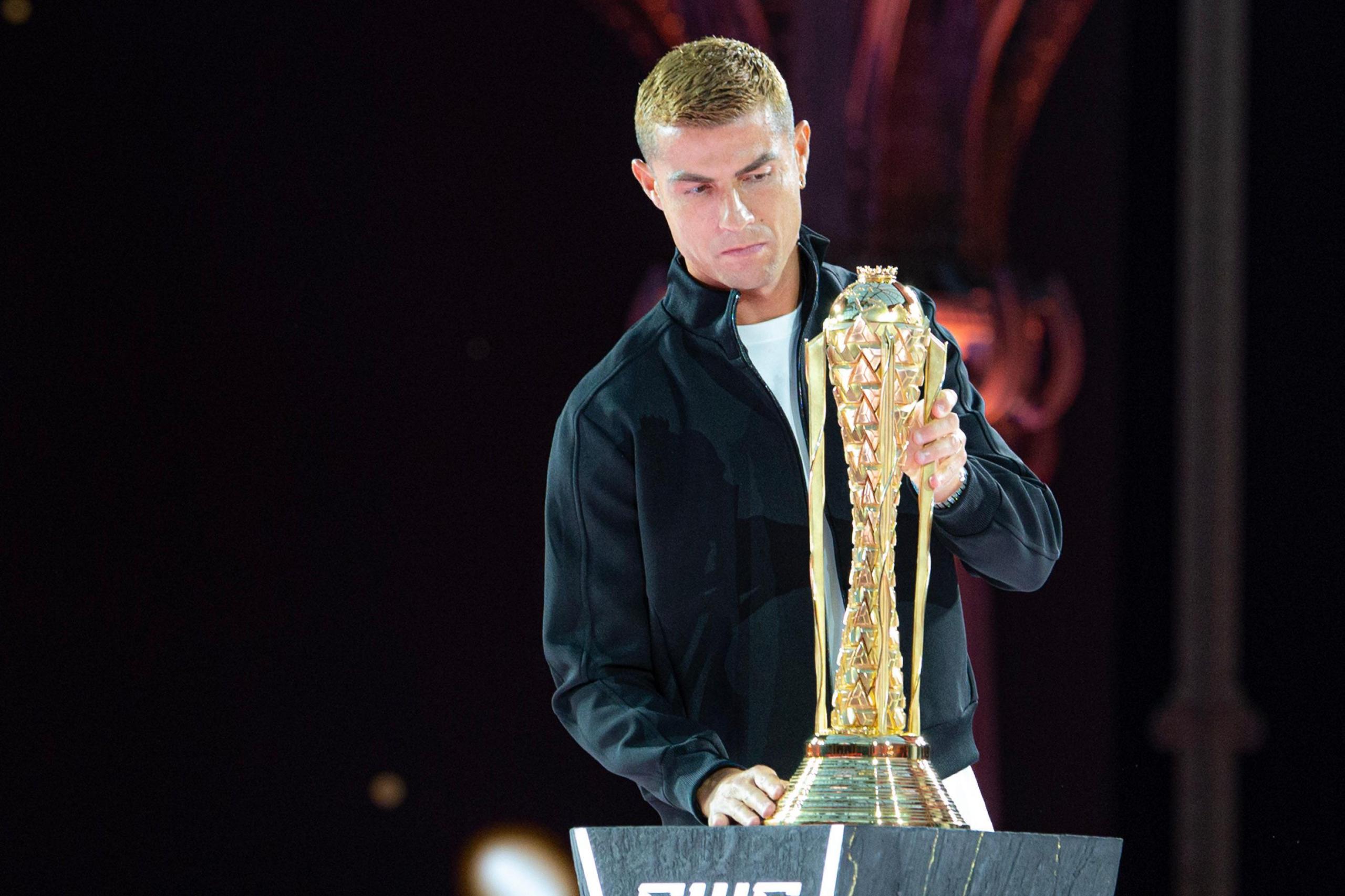 Cristiano Ronaldo, sporting a blonde dyed hairstyle and wearing a black Harrington jacket over a white t-shirt, carefully places the tall, golden EWC trophy on top of a dark grey plinth.
