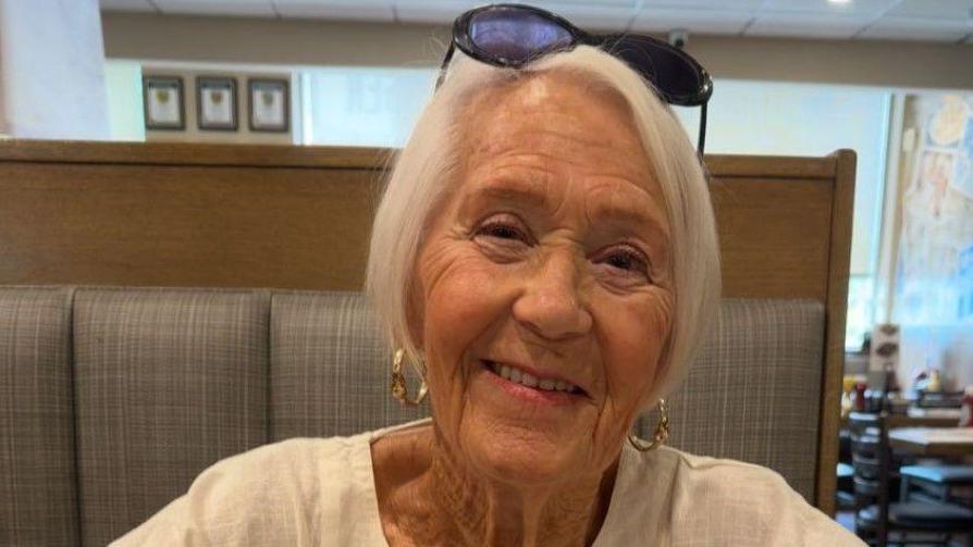 Gloria Stephenson smiling into the camera. She has short, white hair and is wearing light pink lipstick, gold hoop earrings and a white top. She looks to be sitting in a restaurant.