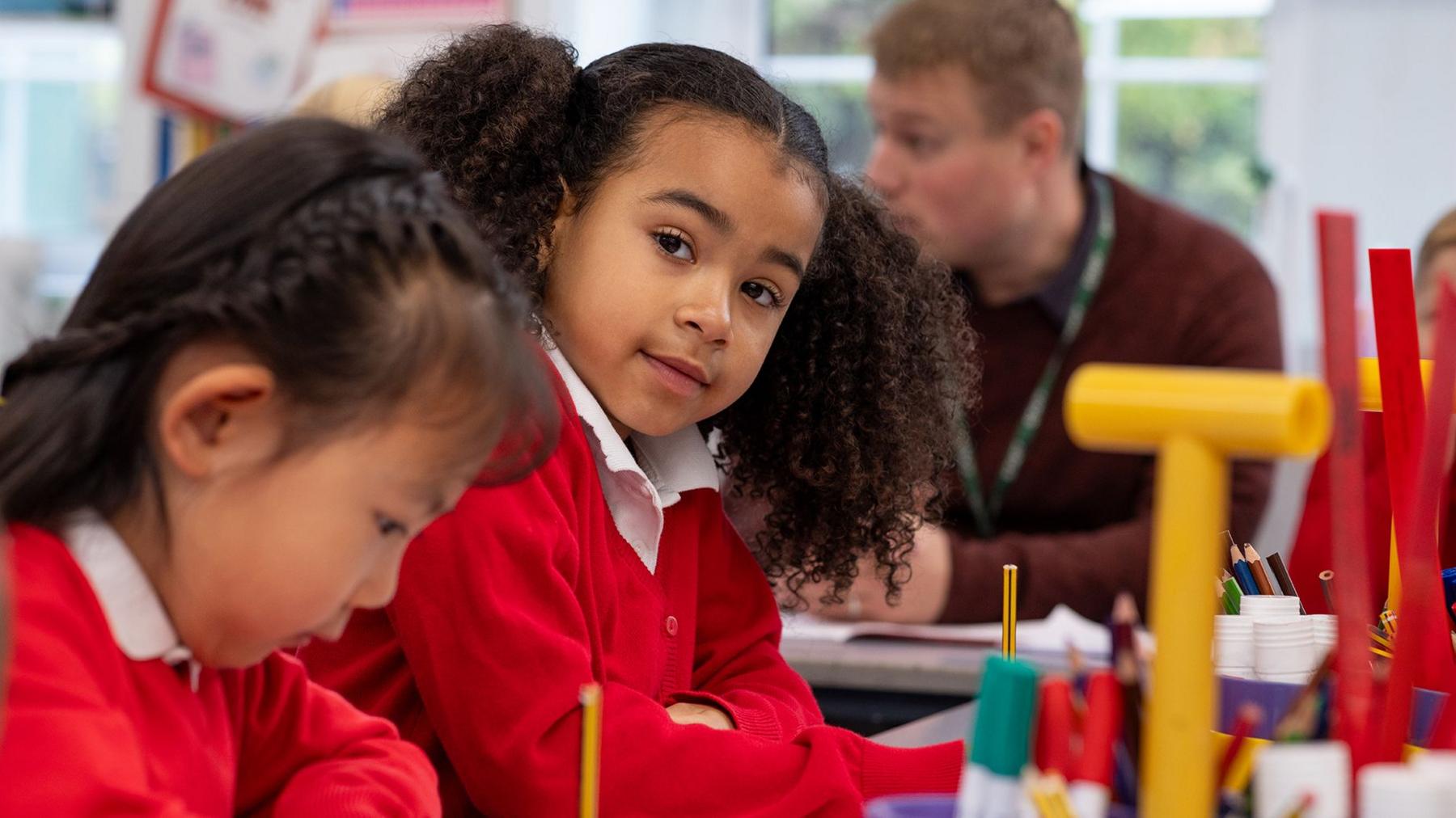 A primary school aged  girl in a red school uniform looks at the  camera as she sits in class. Another student, also a girl in seen in the foreground looking down. A teacher talks to someone else in the background.