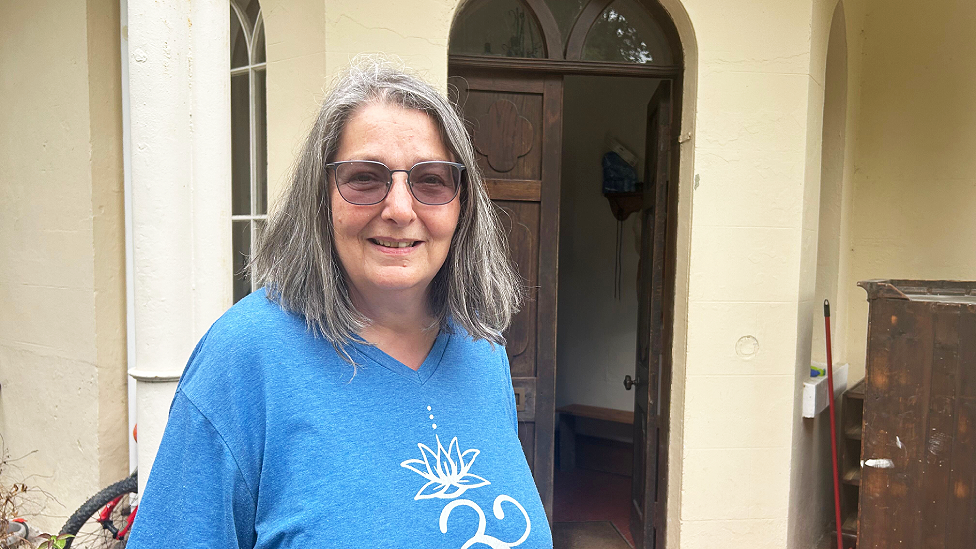 Louise Nicholson is a middle-aged white woman with shoulder length dark grey hair. She wears grey framed photo-reactive glasses, a light blue v-neck t-shirt with a lily pad white emblem on the front. She is smiling at the camera. Behind her is the main entrance to Dol-llys Hall. There are two dark wooden doors with arched fanlights above which open from the middle of a curved porch-type entrance. The building is painted yellowy-cream. There are arched windows to the left of the door visible. A mop handle and a piece of wooden furniture can be seen to the right of the doors.