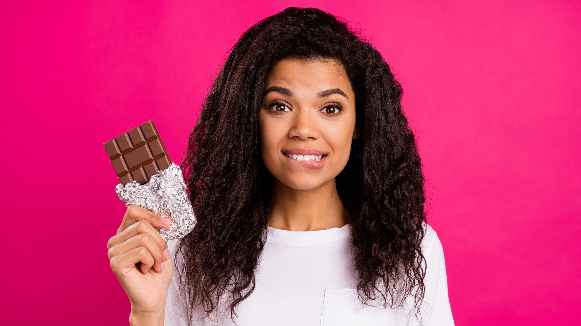 Woman holding a bar of chocolate, she has long black hair and is wearing a white t-shirt and it standing in front of a bright pink background