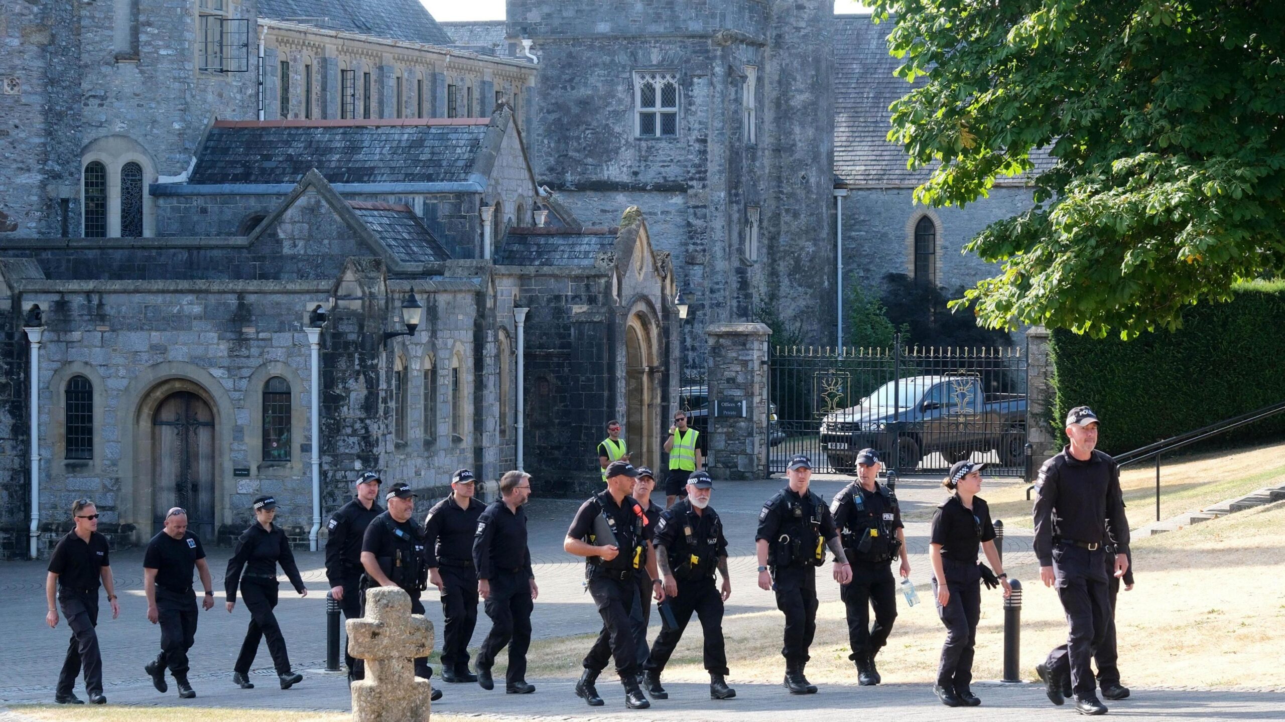 About 14 police officers wearing dark clothing and police caps walking outside on the grounds of Buckfast Abbey.