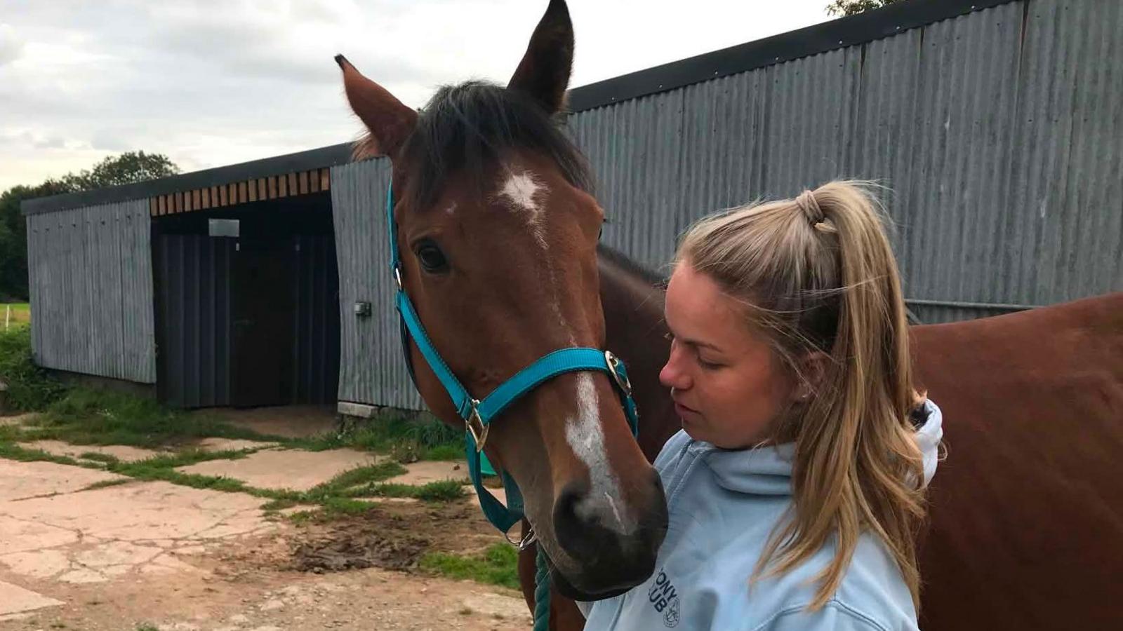 A blonde white woman with her hair in a ponytail wearing with a grey hooded sweater holds a brown horse with a white patch on its nose close to her. Behind them is a yard with a grey corrugated building with a black door