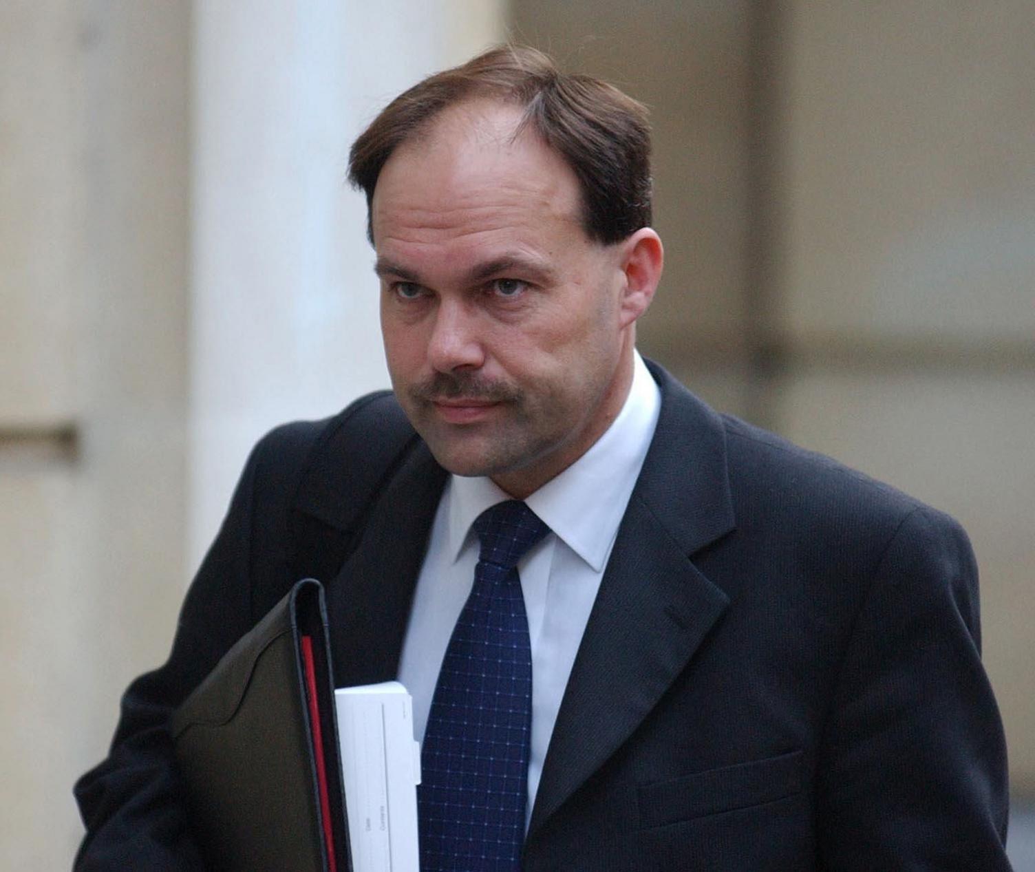 A man with thinning hair and a moustache is walking outside a court building. He is wearing a black suit and tie with a white shirt. He is holding papers contained in a black leather folder.