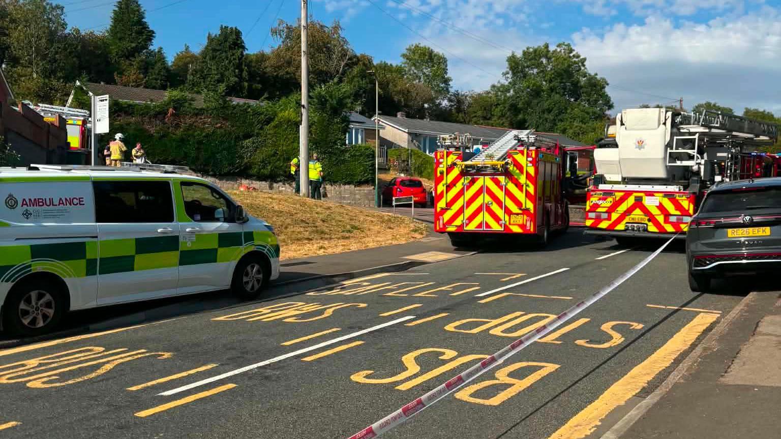 An ambulance van and two fire trucks on a street cordoned off by red and white striped tape. It is a blue sky sunny day and some firefighters can be seen in on the street in the distance