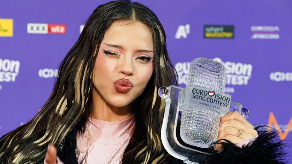 Dara, in a pink top and with blonde streaks in long hair, holds the Eurovision trophy