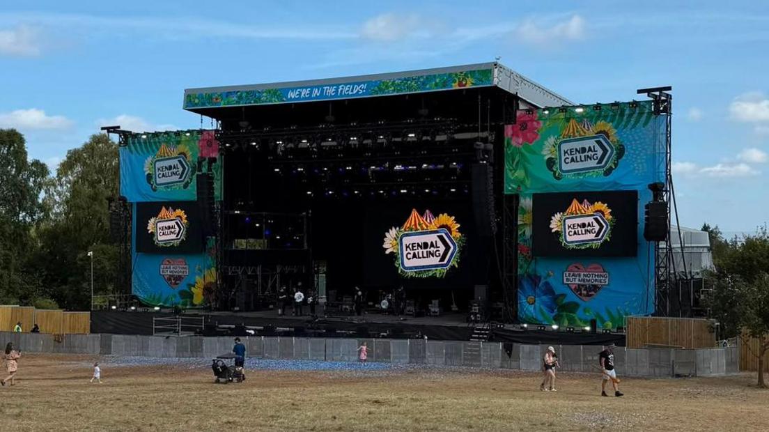 An empty stage at Kendal Calling, with the festival branding. There are a handful of people in the field in front of the stage milling around.