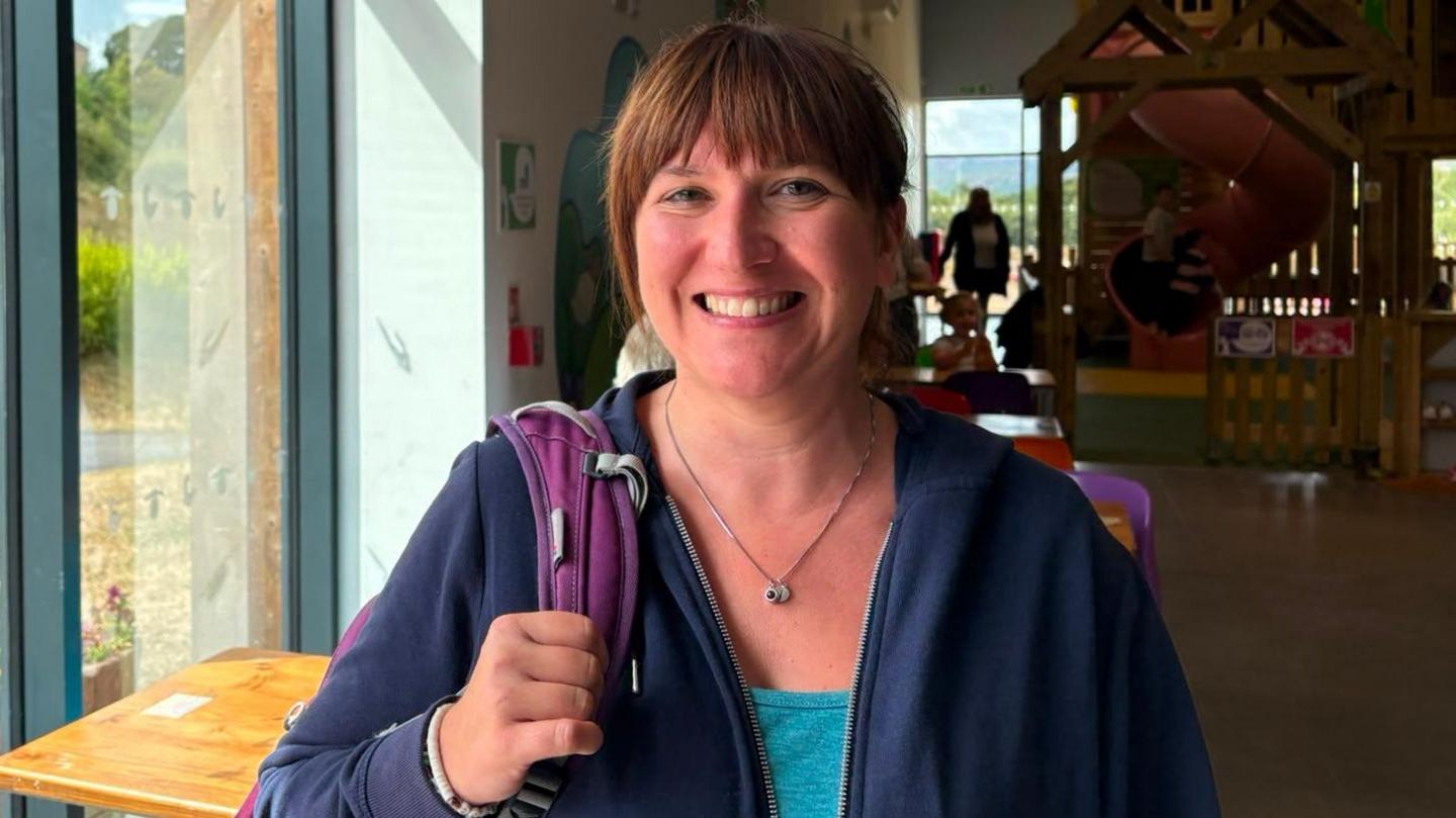 A woman with tied-back brown hair wearing a blue t shirt andf hoodie and carrying a purple backpack. She's smiling and looking at the camera.