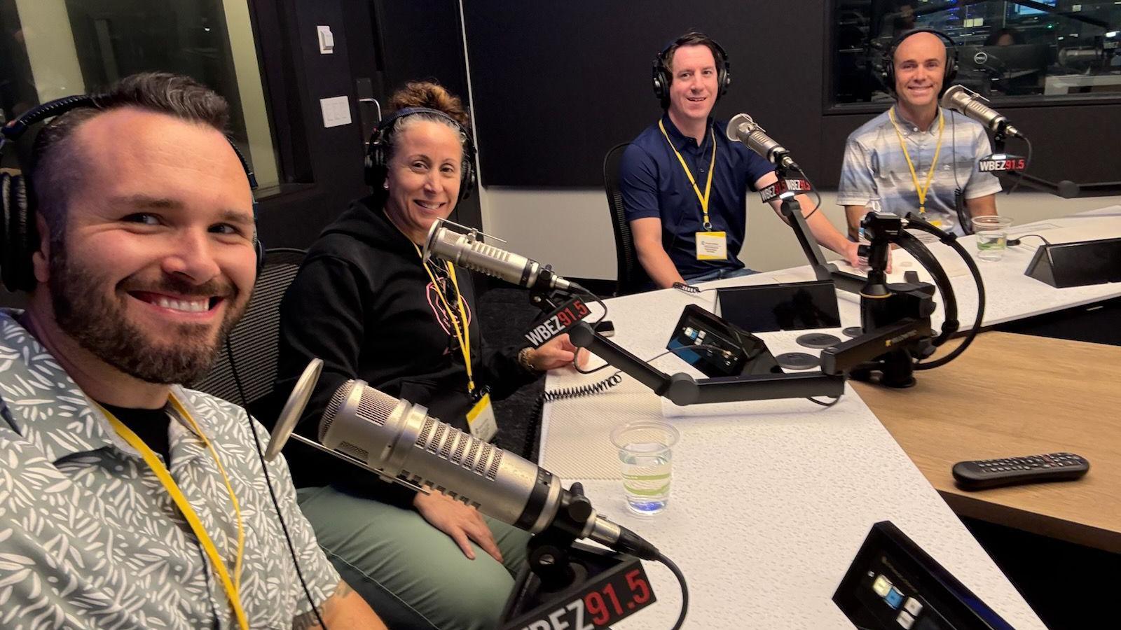 Three men and one woman, all wearing headphones, sit in a podcast recording studio with microphones and other equipment in front of them