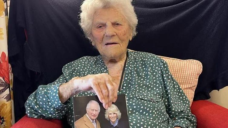 Ethel Caterham sat holding a card with a picture of the King and Queen on the cover.