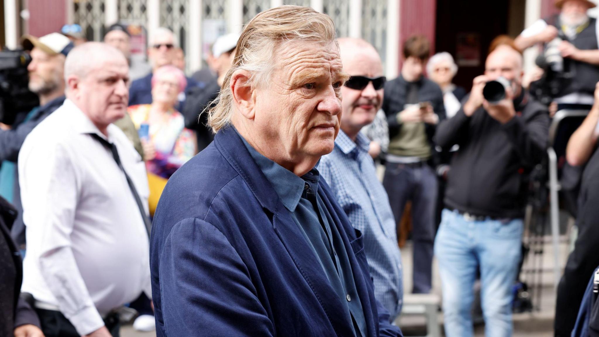 A man wearing a navy suit and navy shirt hold his hands around the front of his waist. He has medium length red hair as he stands in front of a crowd of people, some of whom have cameras.