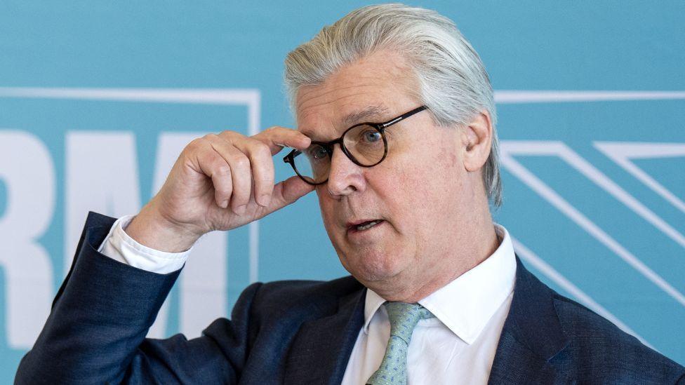 Malcolm Offord, who has grey hair combed back and dark-rimmed glasses, adjusts his spectacles with his right hand while sanding in front of a pale blue branding. He is wearing a dark suit, white shirt and pale blue tie.