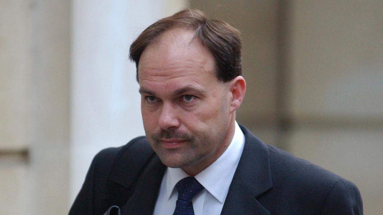 A man with brown hair and a moustache who is wearing a black suit and tie with a white shirt