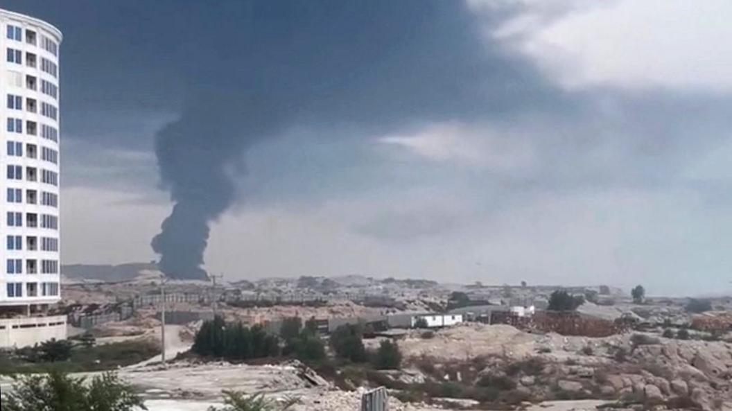 Smoke rises in the distance over a view of a desert-like plane against a cloudy grey sky with buildings in the foreground, in a grab from video showing a strike on Qeshm on 30 July