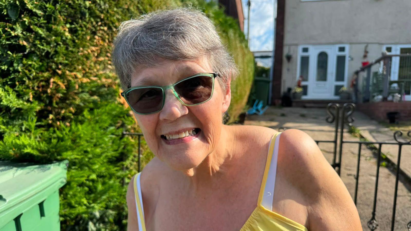 A woman in a yellow dress smiles for the camera. She has short grey hair and stands in front of a black iron gate with a home and hedges also in the background.