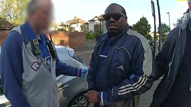 Bodycam footage showing Simon Levy in a navy blue Adidas jacket and sunglasses being detained by police officers on a suburban street. An officer on the left, who's face is blurred, wearing a blue shirt and grey jacket, fitted with a body-worn camera and radio, holds the man's arm.