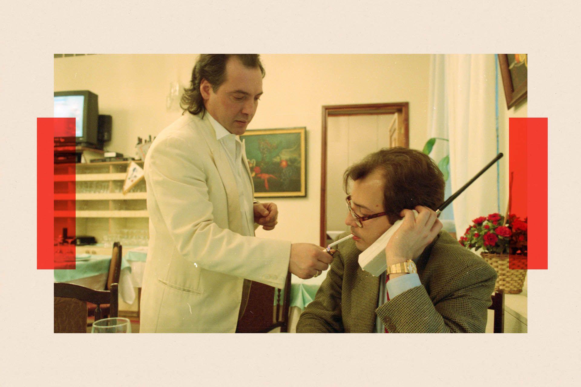 A waiter in a white jacket lights the cigarette of a man sitting at a table holding a large old-style mobile phone
