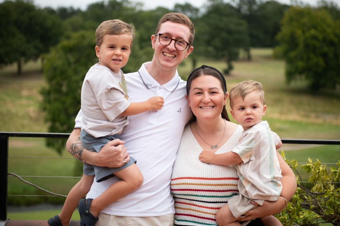 Four people standing together outdoors in the foreground, with two adults in the centre and a child held on each side. A man wears glasses and a white polo shirt, while a woman wears a sleeveless striped top. A metal railing runs behind the group, with grass, trees and open parkland visible in the background.