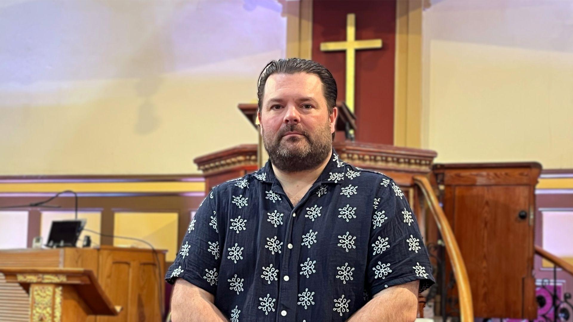 Jonathan stands inside a church in front of a wooden pulpit with a gold cross on a wooden backdrop.