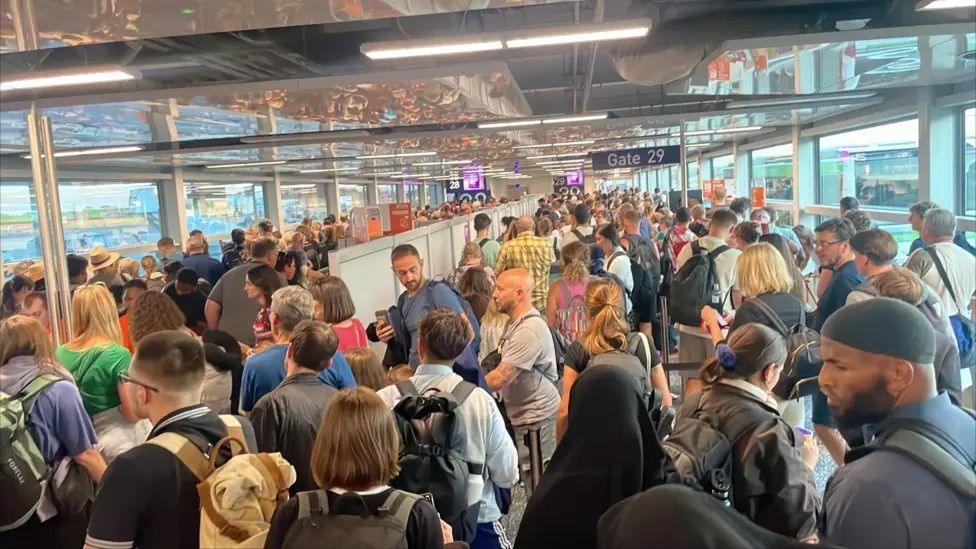 A crowd of frustrated and tired looking passengers pack the halls of a departure gate.