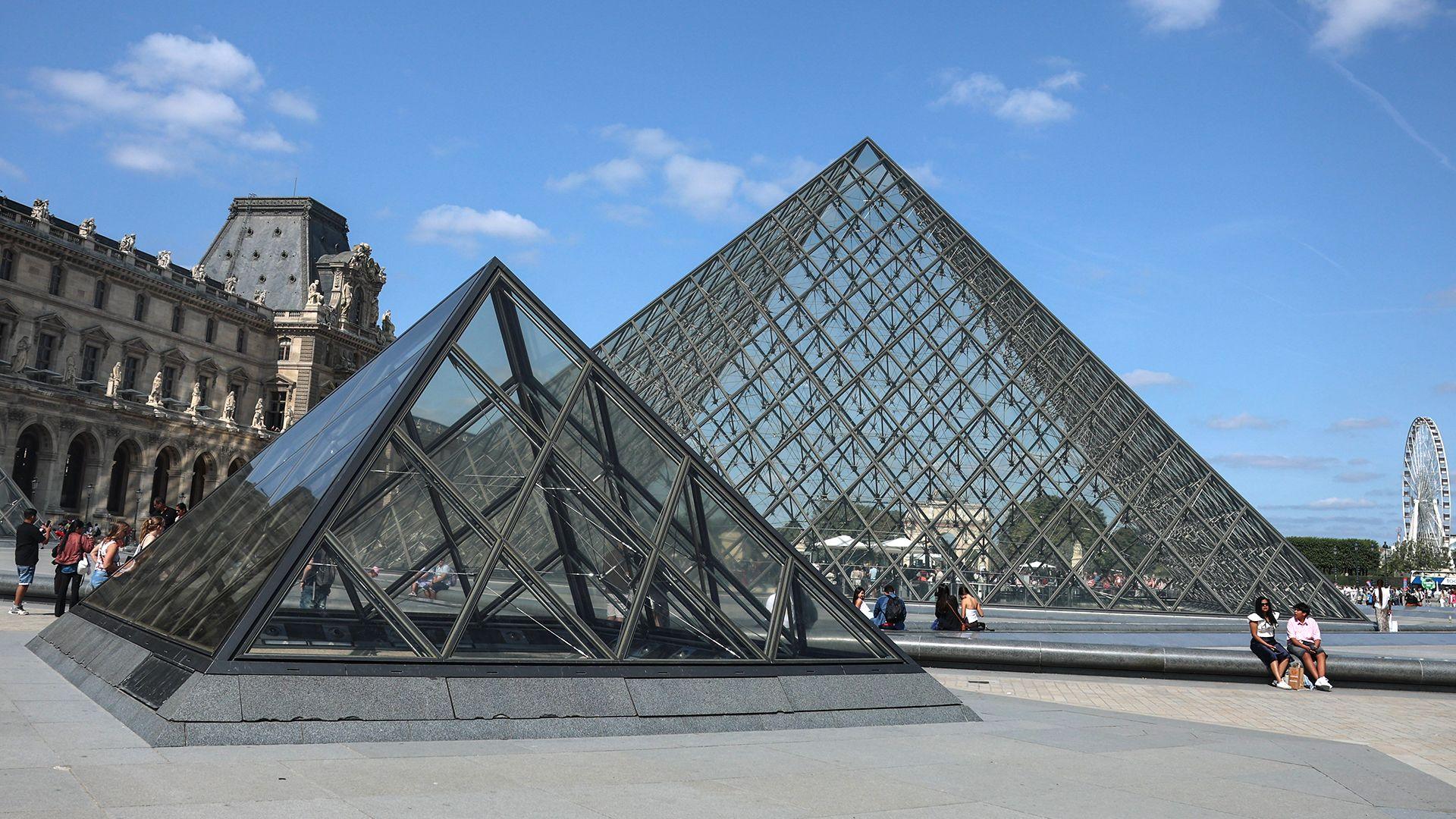 Two modern glass pyramids at the Louvre in Paris with an older building to the left. The sky is blue and there are a few people sitting in front of the pyramids. There is a ferris wheel in the distance on the right.