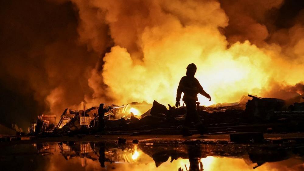 Firefighters work at a site of a Russian missile strike, their silhouettes contrasted against the glowing yellow and orange of smoke plumes illuminated by large fires.