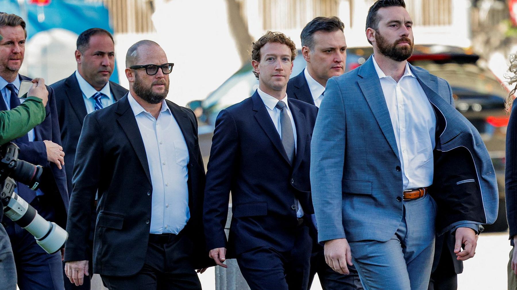 Mark Zuckerberg wearing a suit, looks towards the camera with a blank expression. He is flanked by staff also in suits. Photographed in February 2026