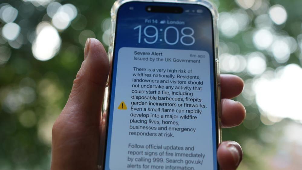 A hand holding an iPhone which includes a notification of an emergency alert from the UK Government on wildfires.
