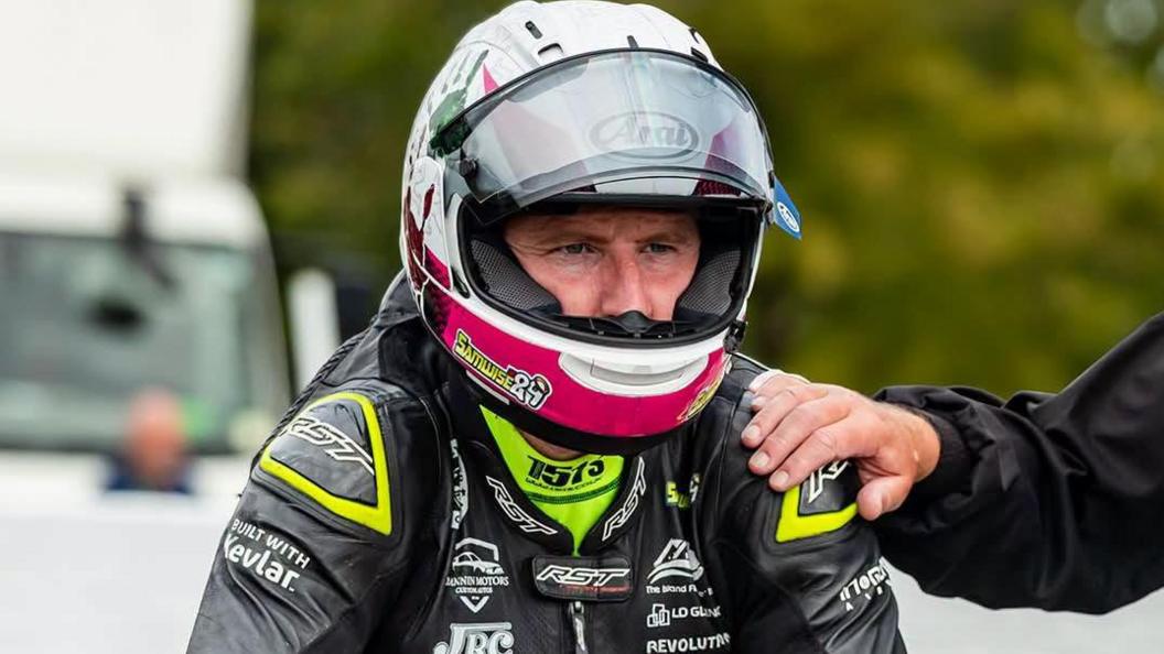 Sam Naughton in his racing leathers and helmet on the start line with a hand on his shoulder.