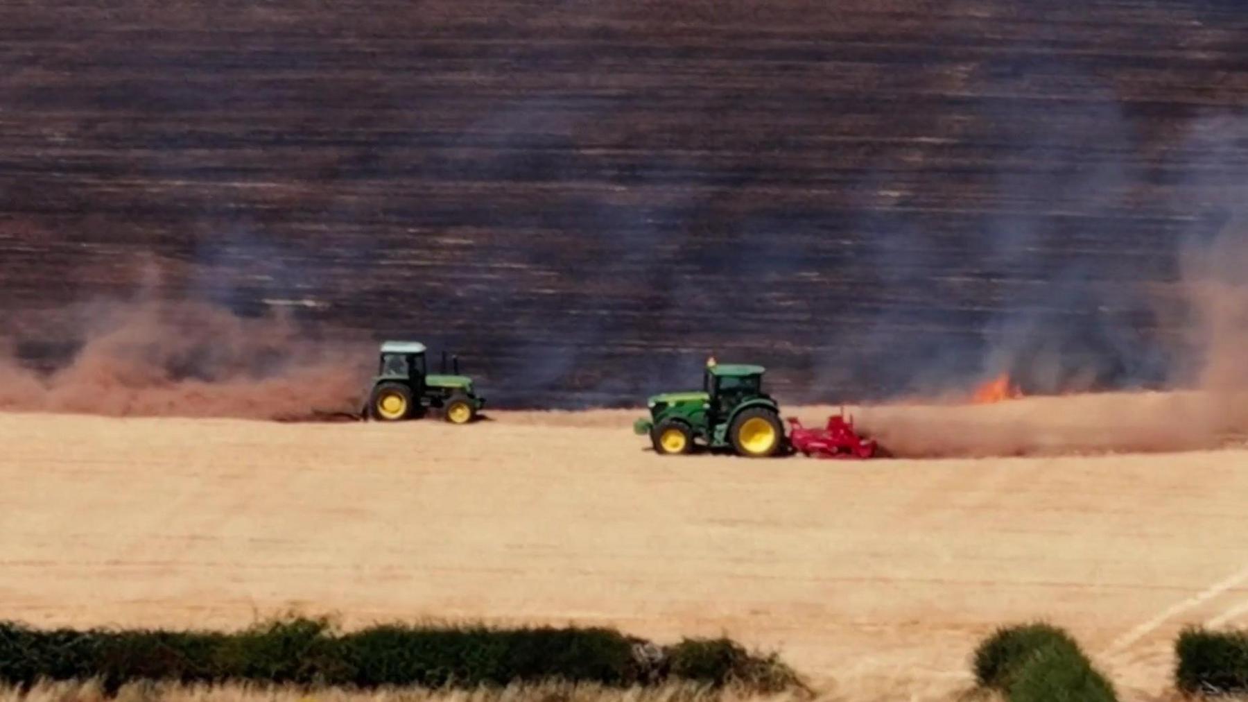 Farmers in green tractors create firebreaks to stop the spread of flames in a field.