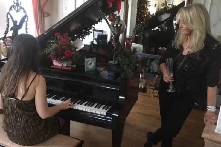 A young girl sat at a black baby grand piano with her back to the camera and Bonnie Tyler standing beside her.