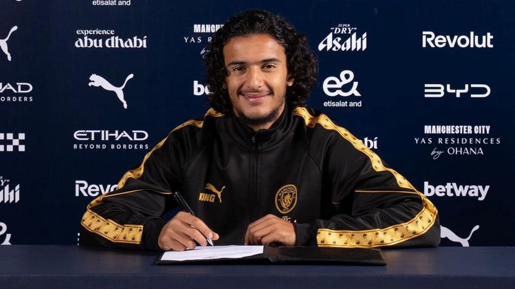 Ayyoub Bouaddi in a Manchester City tracksuit top holding a pen with a contract on the desk in front of him