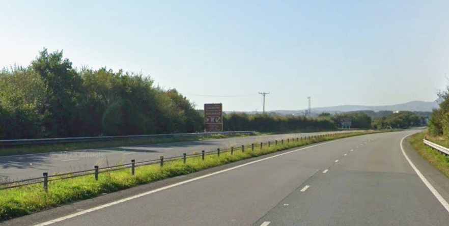 A dual carriageway with green bushes on either side and hills in the background.