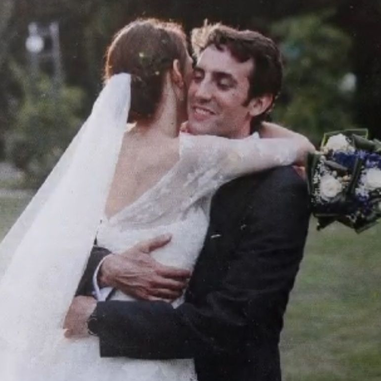 A married couple embrace in a grassy area like a garden. The bride, her back to the camera and wearing a white wedding dress with a long white veil, is being lifted and embraced by the groom. He is wearing a dark suit. She is holding bouquet of white and blue flowers.
