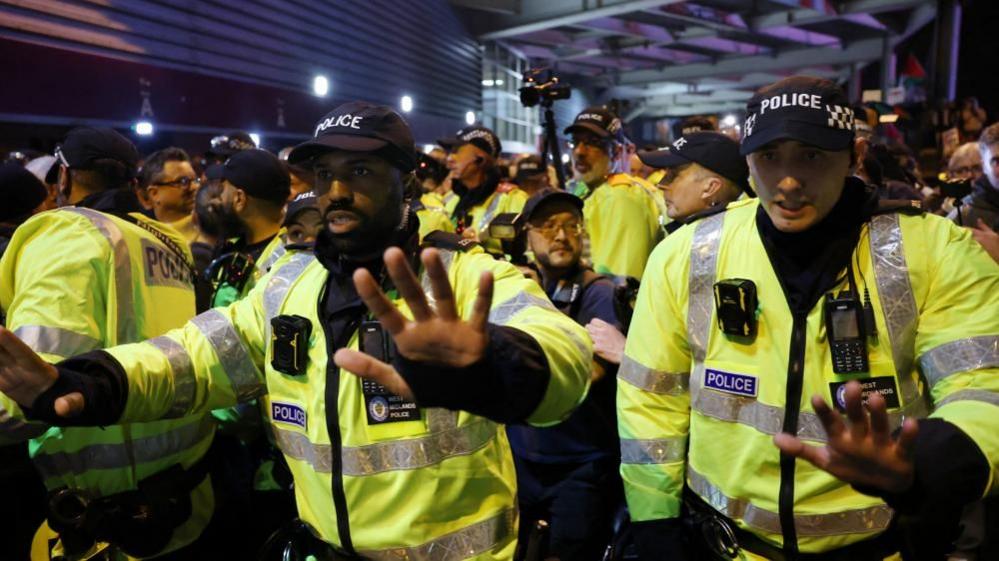 A group of male officers gather outside the stadium. Two of them, one a black man with a moustache and the other white, both wearing caps, have their hands outstretched pushing people back