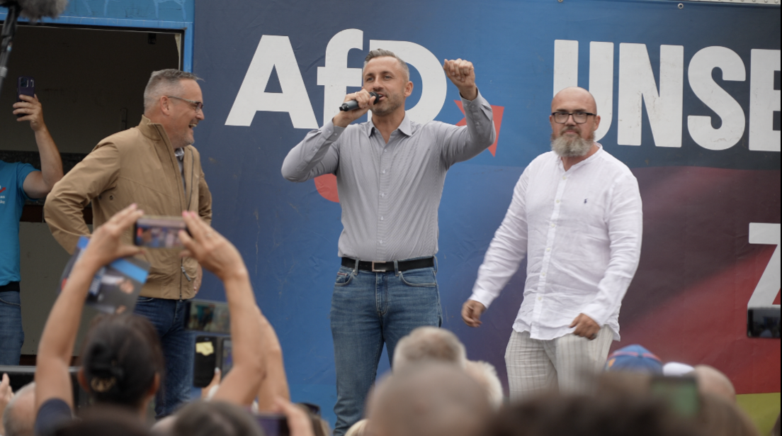Three men are on-stage at an AfD rally. Ulrich Siegmund is in the centre holding a microphone. Crowds are listening and filming on their phones.