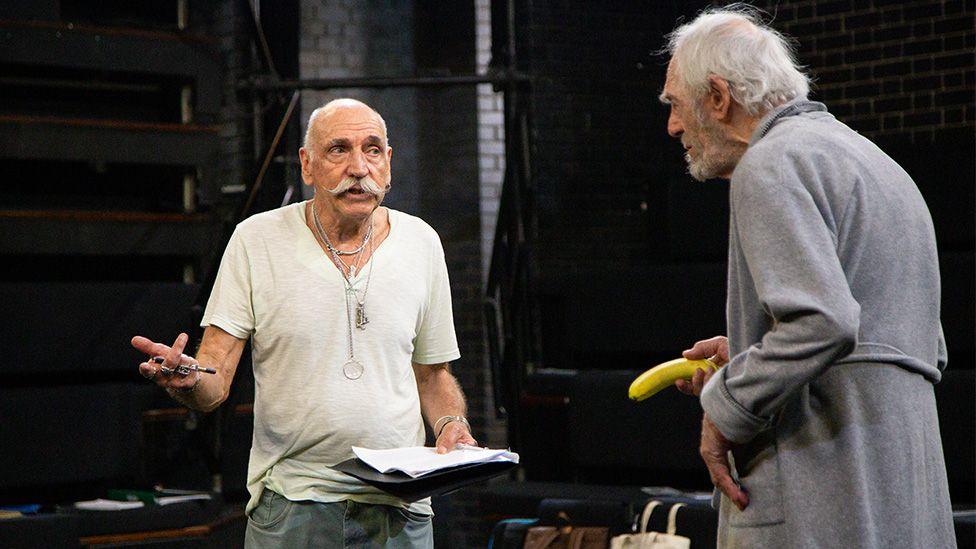Dave Wybrow, with white hair and a handlebar moustache, is wearing a white t-shirt and talking, on stage, to Peter Marinker who is wearing a grey dressing gown and holding a banana as a prop.