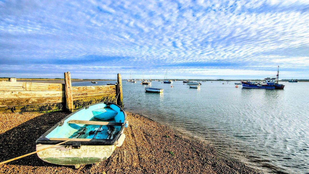 A rowing boat on a shore in the foreground in front of a calm estuary dotted with small boats, under a fairweather sky