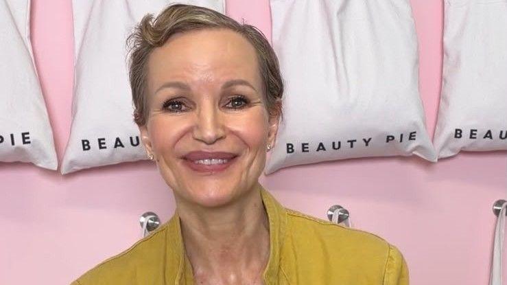 Marcia Kilgore with cropped short brown hair smiling in front of a row of white bag with the Beauty Pie logo on thems at the camera with a blurred background wearing a nude coloured round neck top