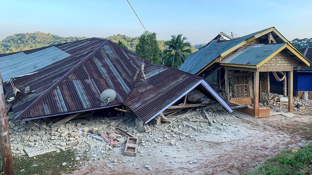 A house has collapsed so that only the roof is visible, lying on the ground.