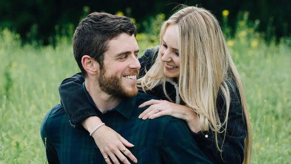 PC Andrew Harper and his wife, Lissie are looking at each other smiling widely. They are in a field which has long reeds of green grass surrounding them. Lissie is hugging PC Harper from behind and leaning over his shoulder to look at him, he is looking over his shoulder at her. PC Harper has short brown hair and a short brown beard and wears a dark green a blue checked shirt. Lissie has long blonde hair and wears a black denim and shearling lined jacket. She is wearing an engagement ring on her ring finger.