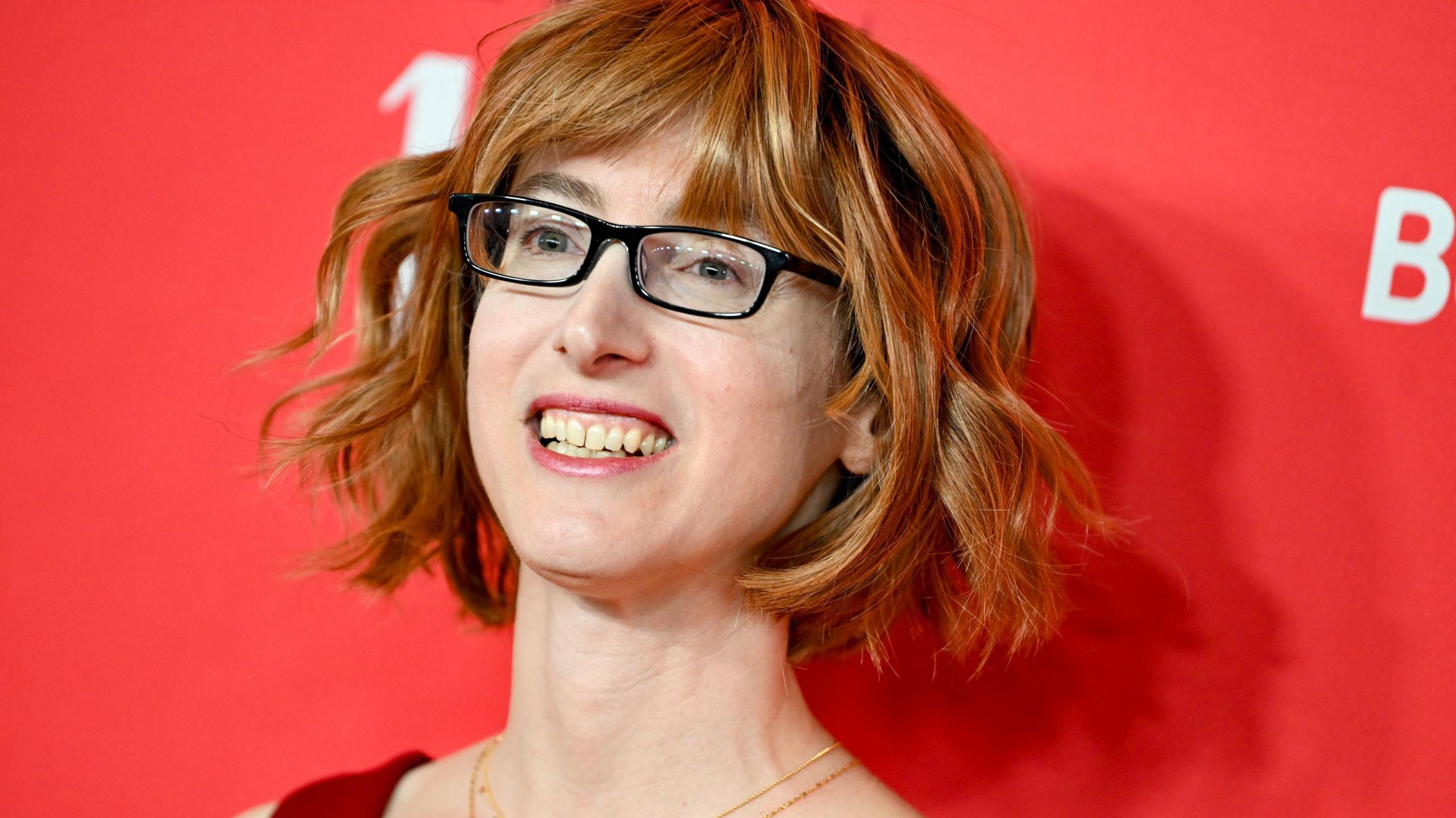 A woman wearing a red dress standing in front of a red backdrop. She is wearing a necklace, glasses and has short wavy hair