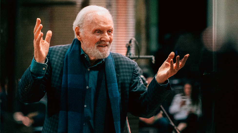 Sir Anthony Hopkins in a music studio. He wears a green and dark blue checked blazer and a blue plaid scarf, with a denim shirt underneath. He has both hands in the air and smiles.
