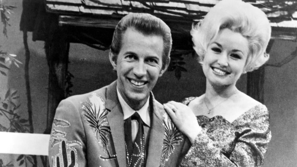 Wagner sits on a stool in a suit with emblems of cacti and plants in this black and white photogragh. He is posing with his guitar.  Dolly stands behind him leaning on his shoulder.  Her hair is short but still standing on end.