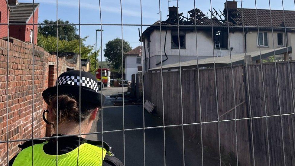 A police officer is stood facing a badly burnt property. The roof has caved in and exposed the wooden beams. An alley runs down next to the property and it has a wooden fence on one side