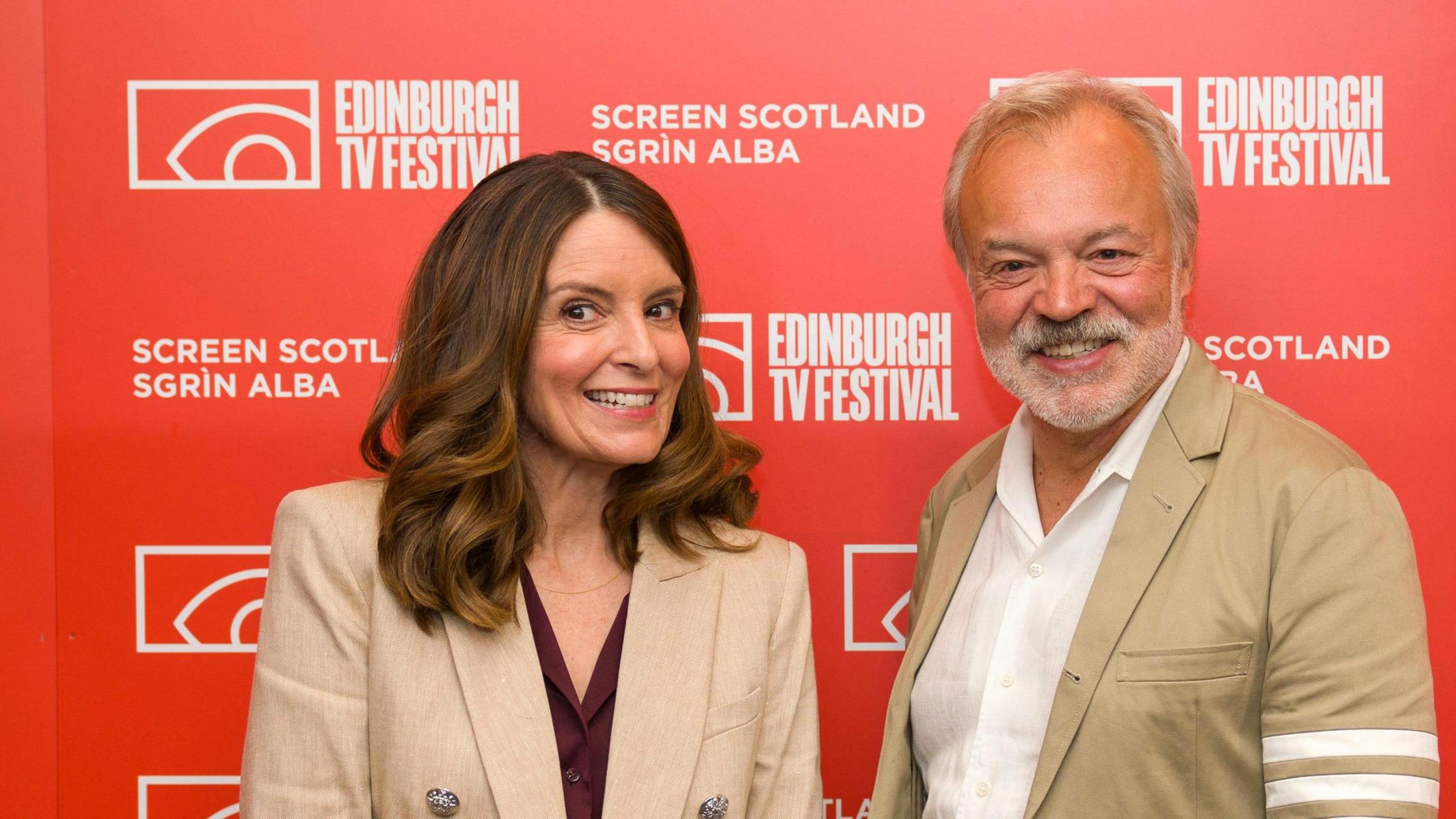 American comedian, writer and producer Tina Fey with presenter Graham Norton at the Edinburgh TV Festival, in the Edinburgh International Conference Centre. They stand in front of a red and white Edinburgh TV Festival backdrop.