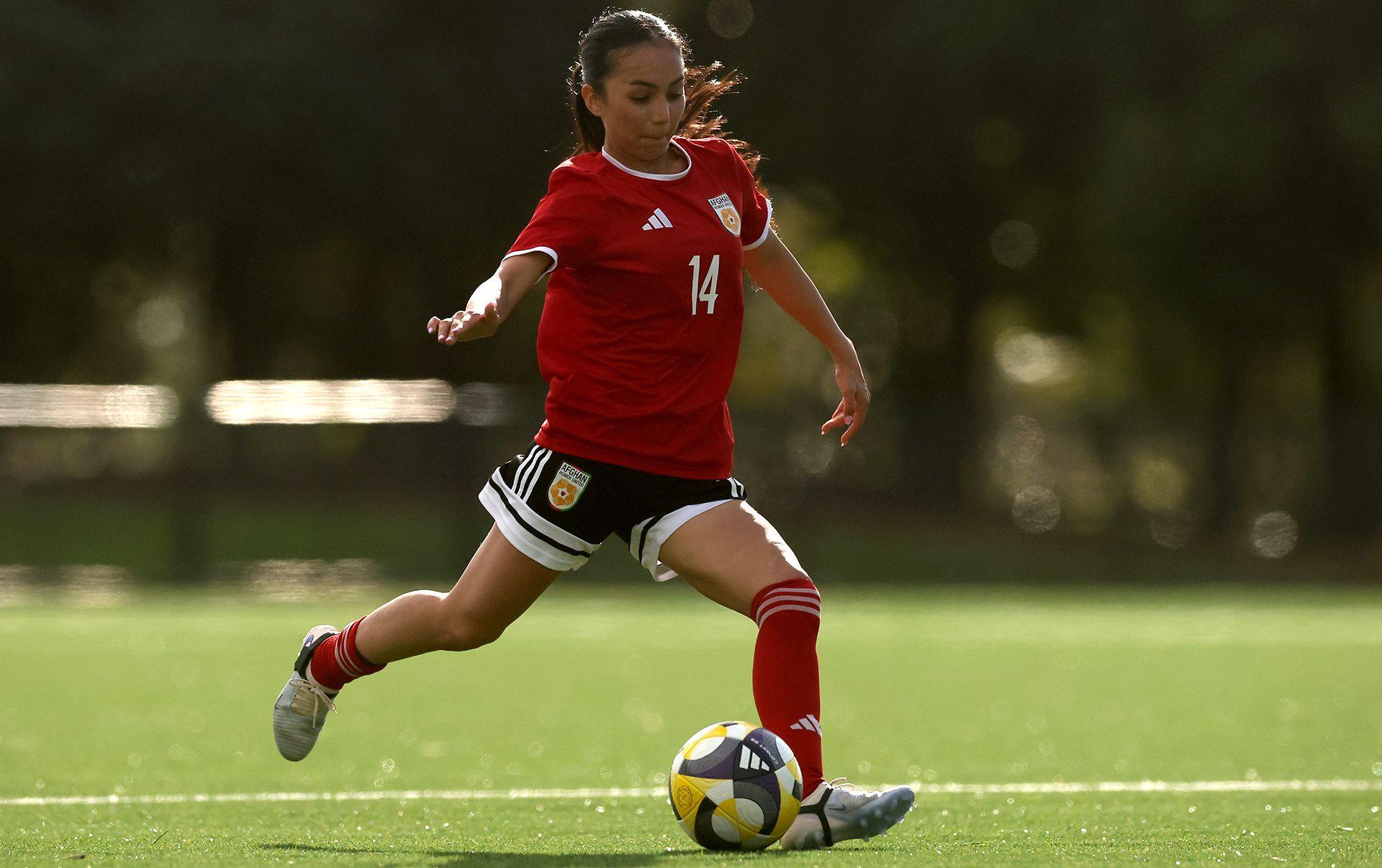 Action shot of a woman about to kick a football. She is wearing a red shirt with the number 14 on it in white. She has black shorts, red socks and white boots. Her long dark hair is tied in a ponytail.