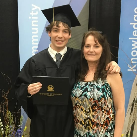 Jared puts his arm around his mother's shoulders and holds a plaque in the other. He wears a graduation cap and gown and has short dark hair. Kelly has long brown hair and wears a multi-coloured patterned dress with blue and orange tones.